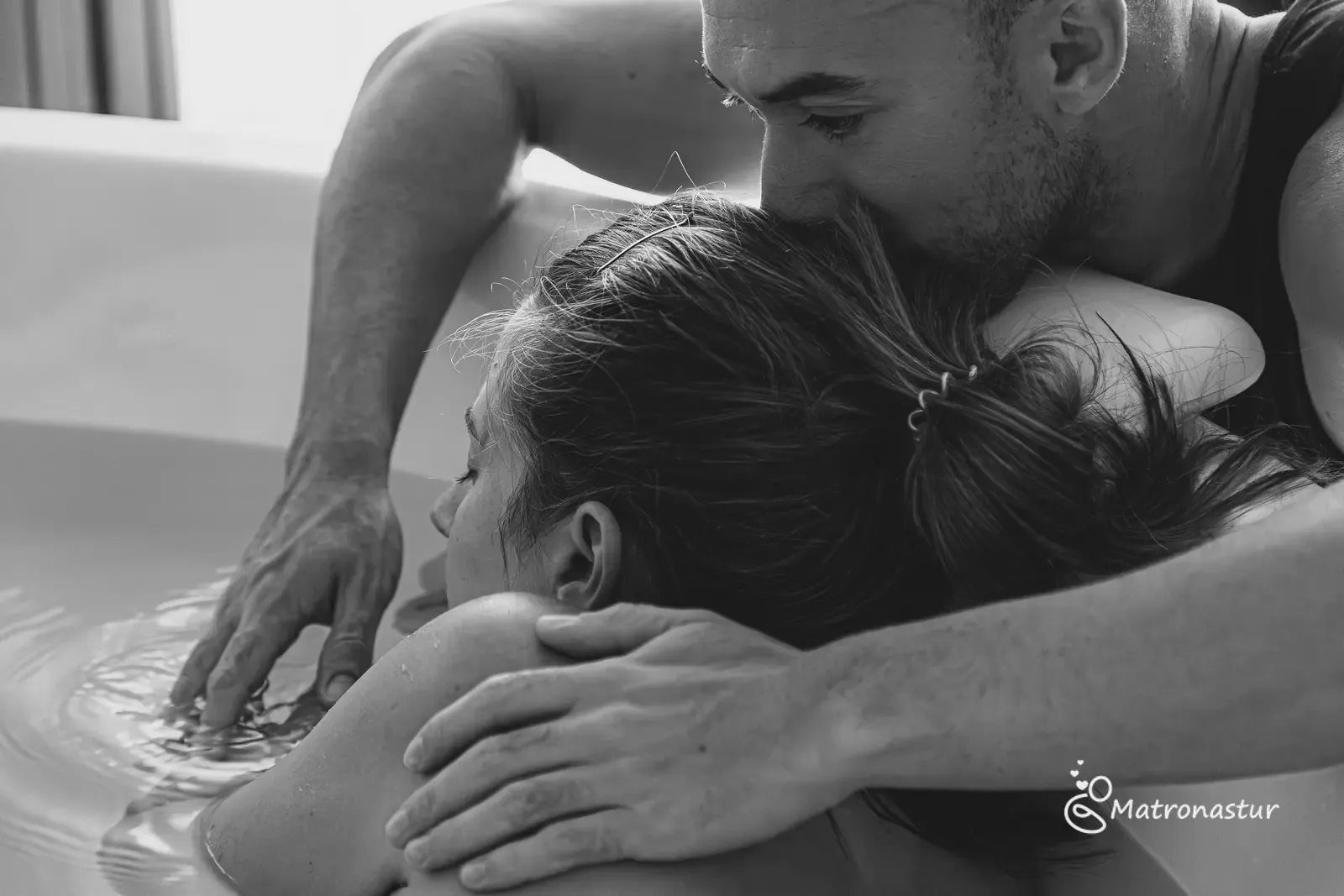 Néstor sostiene a Tania dentro de la bañera de partos y le da un beso en la cabeza, fotografía en blanco y negro
